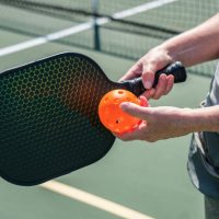 Hand holding a pickleball paddle and ball on a court.