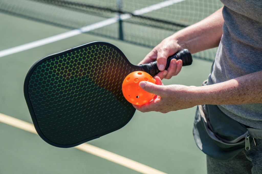 Hand holding a pickleball paddle and ball on a court.