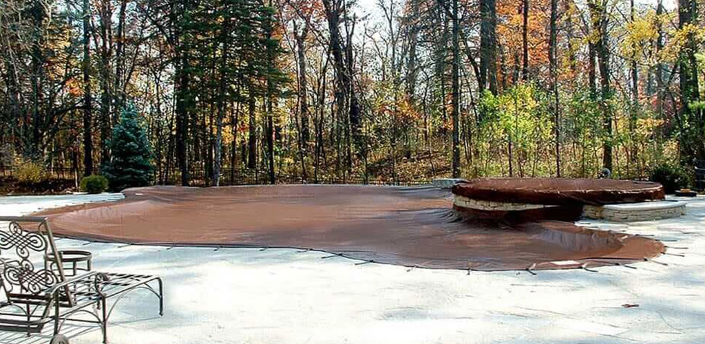 Brown pool cover stretched over an in-ground pool near trees.