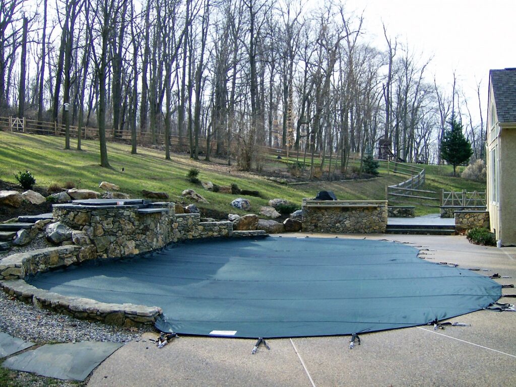 Backyard pool enclosed with a dark gray safety cover near a landscaped yard.
