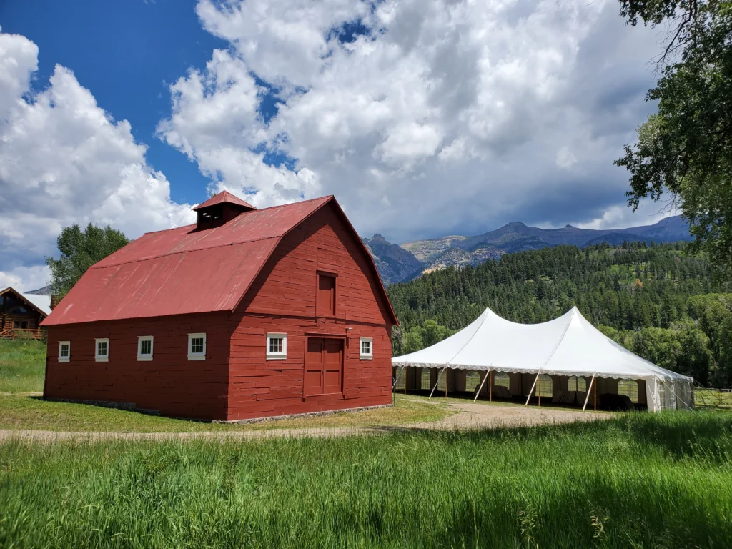 White pole tent by Anchor Industries set up beside red barn in mountain setting