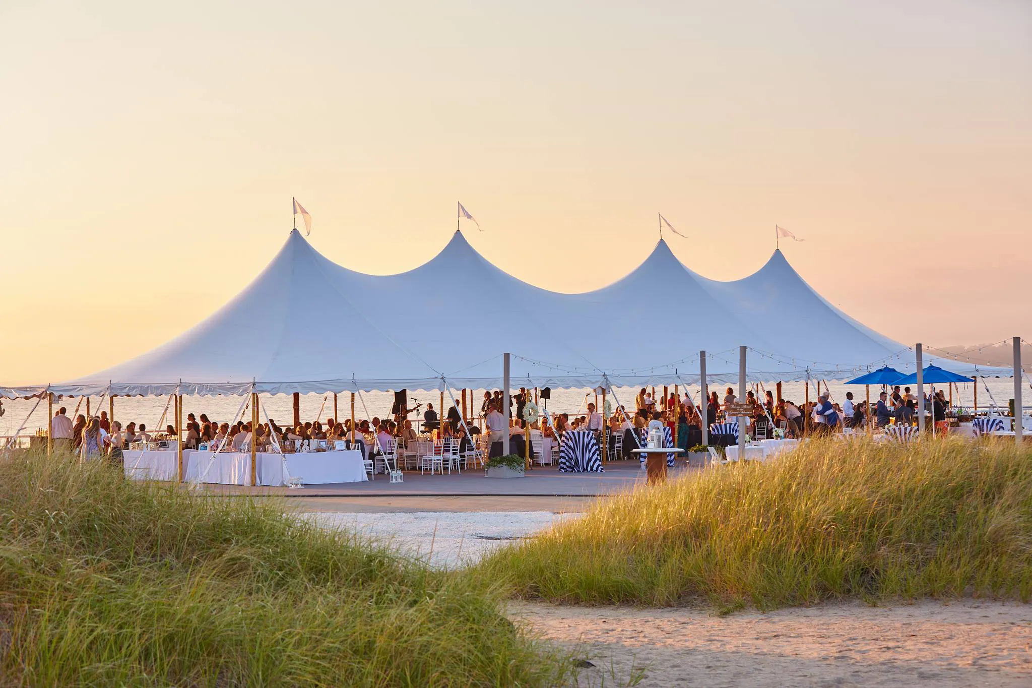 White sailcloth tent near sand dunes and ocean at sunset.